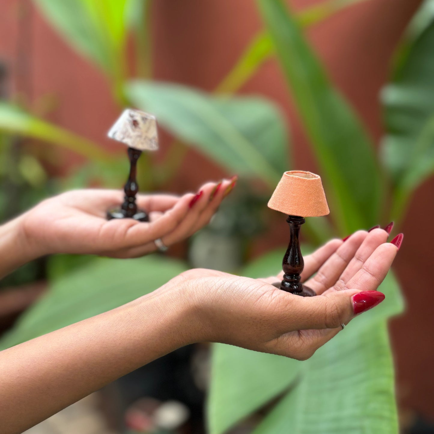Two hands holding small lamp sculptures against a natural background