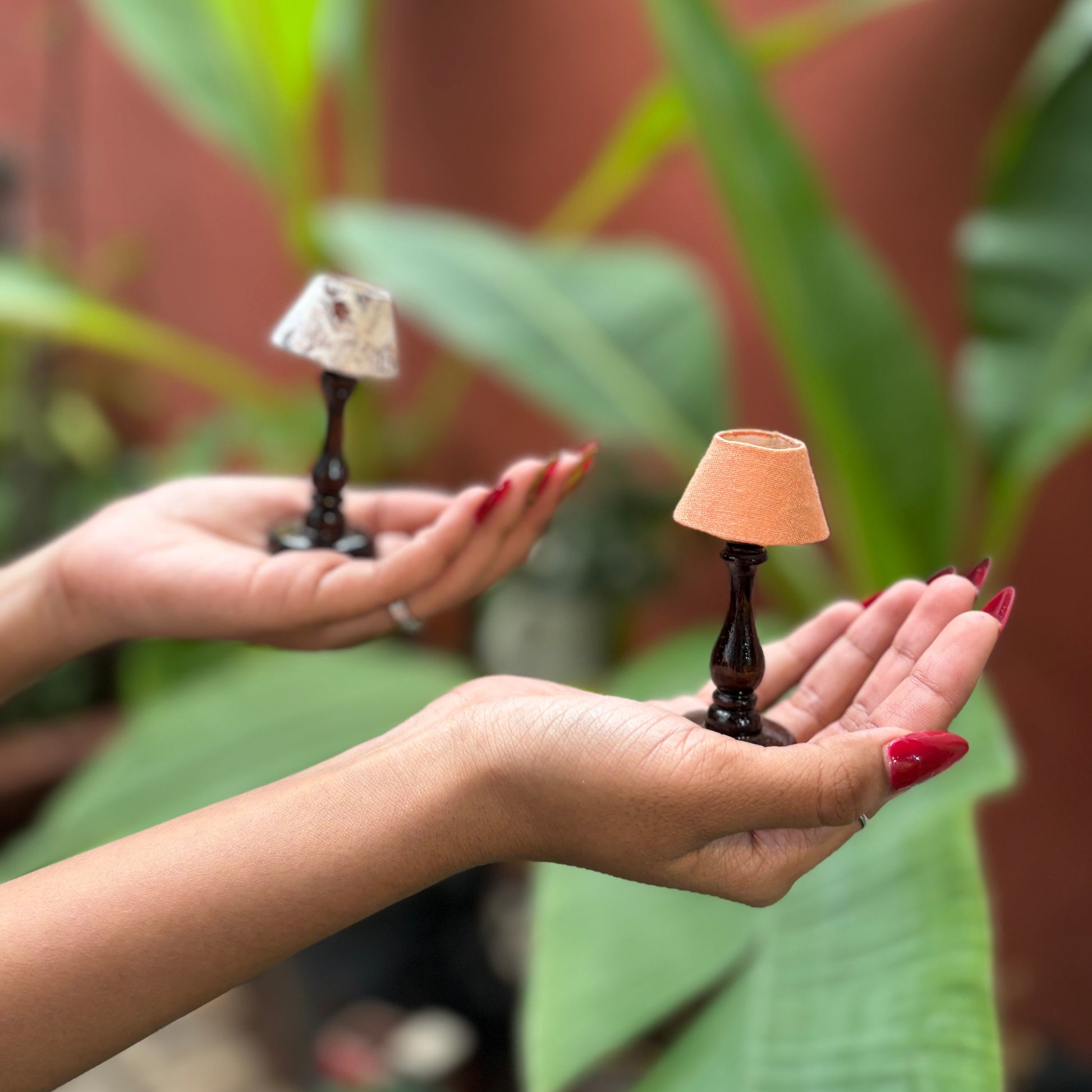 Two hands holding small lamp sculptures against a natural background