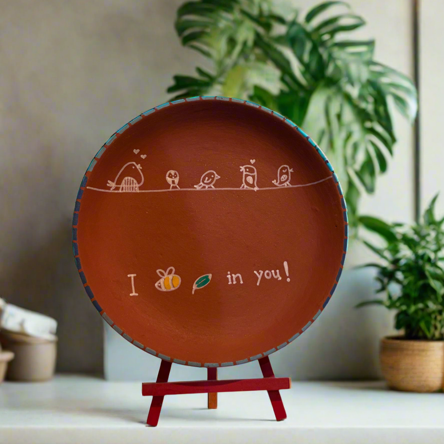 Decorative plate with text and illustrations on a stand, placed on a surface with plants in the background.